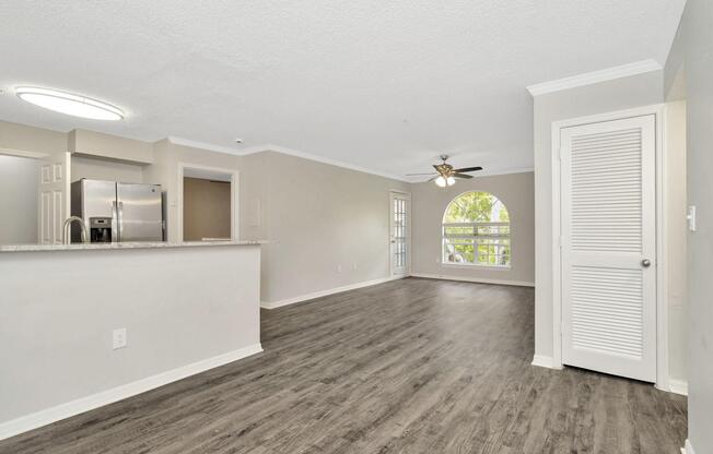Spacious living area featuring a light-filled room with an arched window, modern ceiling fan, and open layout. The space includes a kitchen with a breakfast bar and stainless steel appliances, complemented by neutral walls and wood-like flooring.
