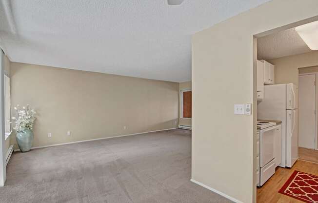 A spacious living room here at Watercrest with soft carpet, neutral walls, and a large window, opening to a kitchen with white appliances and wood-style flooring, creating a bright and open living space.