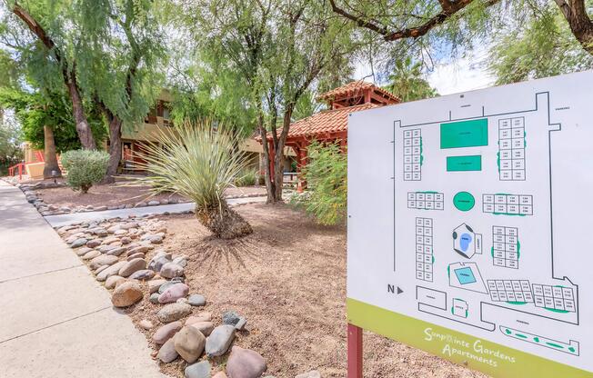 Map showing the layout of Sympatico Gardens Apartments, with nearby landscaping featuring rocks, shrubs, and trees. The two-story building can be seen behind the sign, which also includes a key to different apartment locations and amenities within the complex.