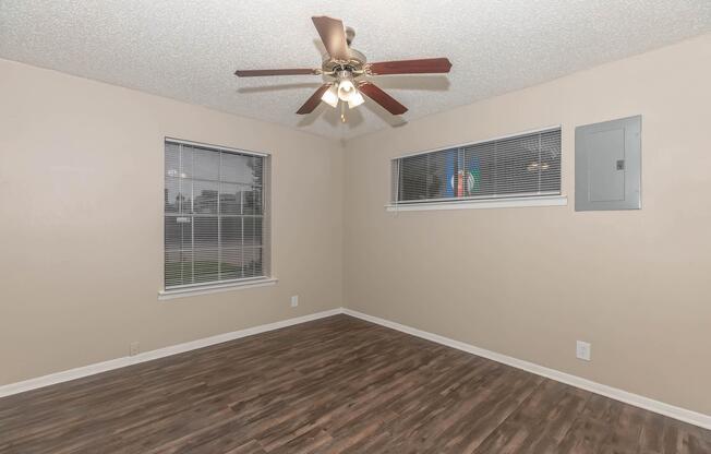 A well-lit, empty room featuring light-colored walls, a ceiling fan with light fixtures, two windows with blinds, and a brown laminate floor. An electrical panel is mounted on one wall. The space is ready for furniture and decoration.