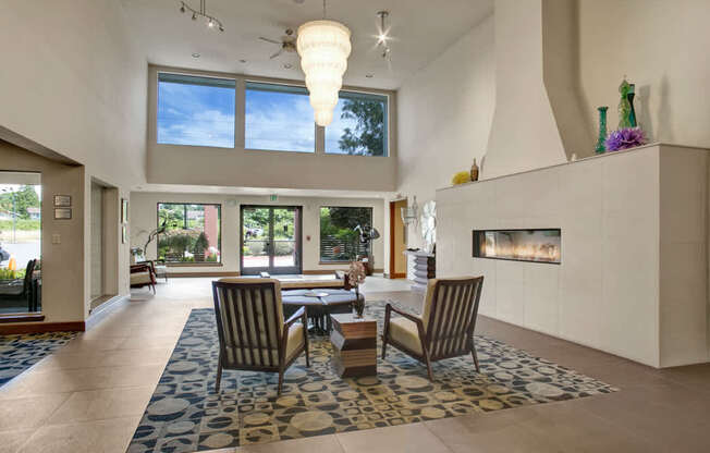 A bright resident lounge here at Heritage Hills featuring soaring ceilings, a dramatic pendant chandelier, and a tiled fireplace wall. The space includes a patterned area rug, comfortable accent chairs arranged around a low table, large windows that bring in natural light, and an open layout that creates an inviting setting for relaxing or socializing.