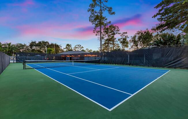 A tennis court with a blue surface and white lines is surrounded by a fence and trees.