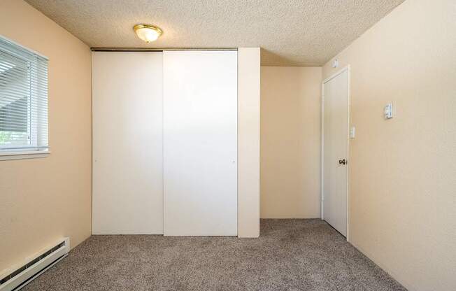 A room with a carpeted floor, a window with blinds, a white door, and a light fixture on the ceiling.