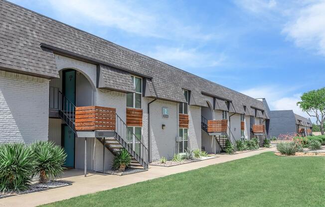 A modern apartment complex exterior featuring a row of two-story buildings with balconies. The buildings have light-colored brick walls, dark gray roofs, and green doors. The surrounding landscape includes well-maintained grass and shrubs, with clear blue skies in the background.