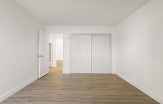 A room with white walls and wooden flooring with a door on the left and a closet on the right.