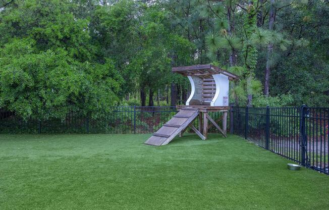 A wooden play structure with a slide and stairs sits on artificial green grass, surrounded by a black fence. Lush green trees and foliage fill the background, creating a serene outdoor environment.