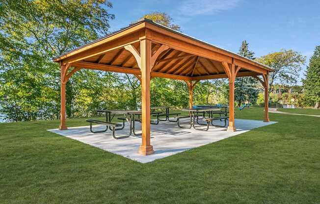 A wooden pavilion with a white base and a roof is surrounded by a grassy area.