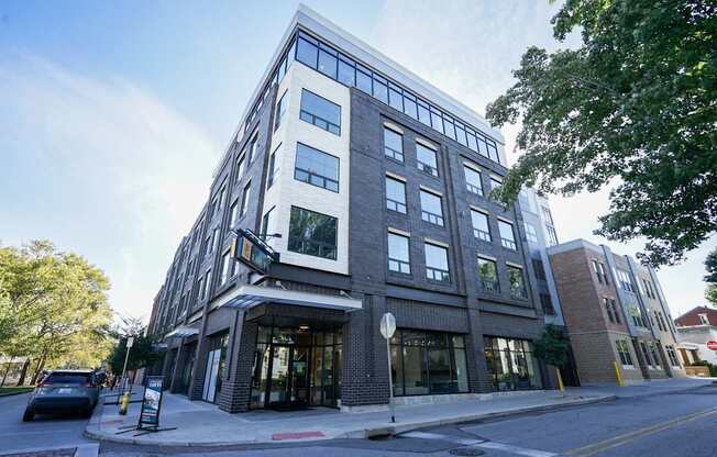 A modern multi-story building with a glass facade and a brick exterior is situated on a street corner with a car parked in front.