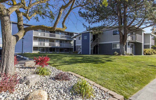 Exterior view of manicured grounds and building. Rock garden, large trees and blue skies in the background at The Lakes Apartments, Moses Lake, WA.