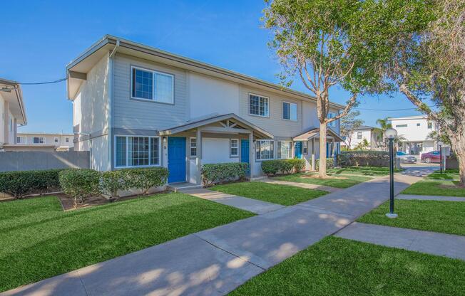A well-maintained two-story apartment building with blue doors, surrounded by neatly trimmed grass and trees. A paved walkway leads to the entrance, with additional residential buildings visible in the background. The scene is bright and inviting, showcasing a pleasant living environment.