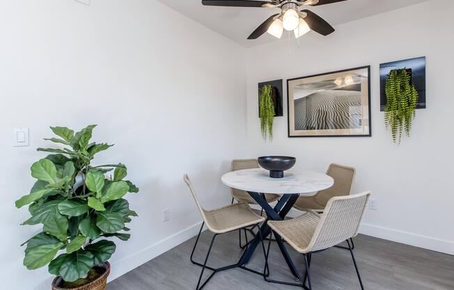 A modern dining area featuring a round marble table with four woven chairs. A large framed photograph hangs on the wall, accompanied by two wall-mounted planters with hanging green plants. A potted plant is positioned in the corner, and a ceiling fan is visible above.