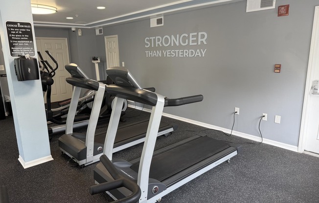 A fitness room featuring two treadmills against a gray wall with the motivational phrase "STRONGER THAN YESTERDAY" prominently displayed. The floor is covered in black rubber flooring, and there are exercise machines in the background, creating an inviting workout environment.