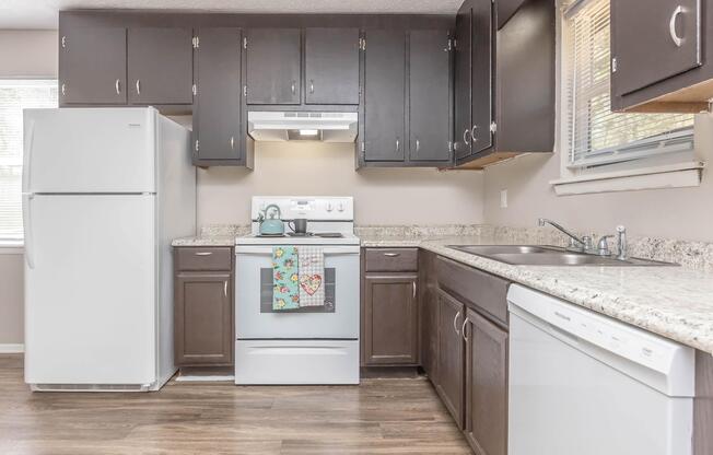A modern kitchen with dark wood cabinets, a white refrigerator, and an electric stove. The countertop is a light granite, and there is a dishwasher beside the sink. A colorful towel hangs from the stove. Natural light streams through a window, illuminating the space.