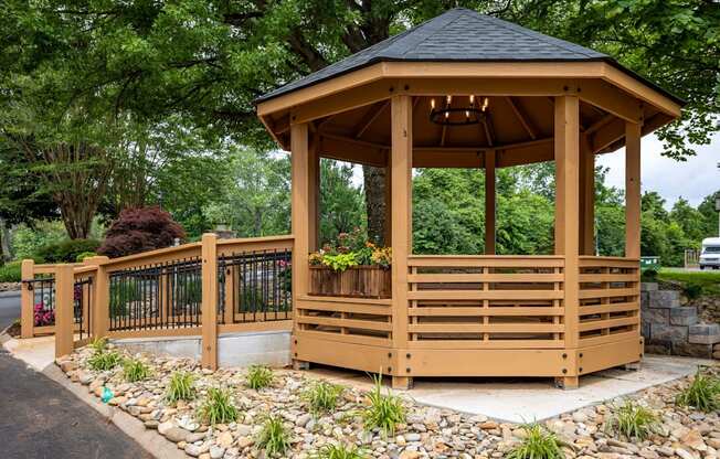 A wooden gazebo with a black roof is surrounded by a metal fence and a gravel area with plants.