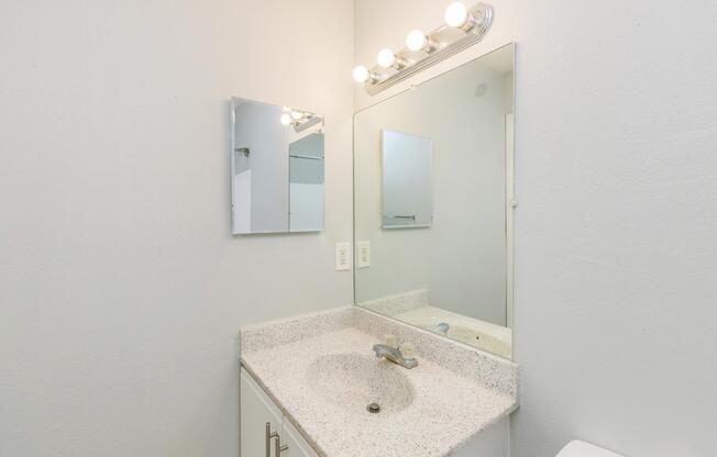 A bright, modern bathroom with a white countertop and sink. Above the sink, there is a rectangular mirror and five light fixtures providing illumination. The walls are painted light gray, creating a clean and airy atmosphere. A towel rack is visible in the background.
