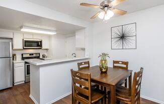 Modern kitchen and dining area featuring a wooden table with four chairs, a ceiling fan, and a decorative vase on the table. The kitchen includes stainless steel appliances, white cabinetry, and a countertop extending into the dining space. A wall art piece adds a contemporary touch.