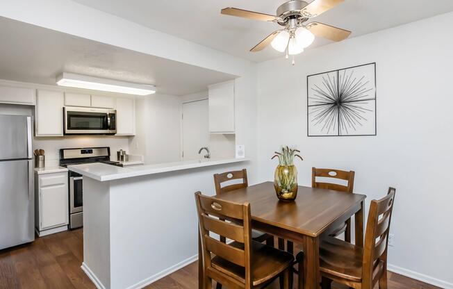 Modern kitchen and dining area featuring a wooden table with four chairs, a ceiling fan, and a decorative vase on the table. The kitchen includes stainless steel appliances, white cabinetry, and a countertop extending into the dining space. A wall art piece adds a contemporary touch.