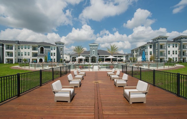 A wooden deck with white chairs and a black fence in front of apartment buildings.