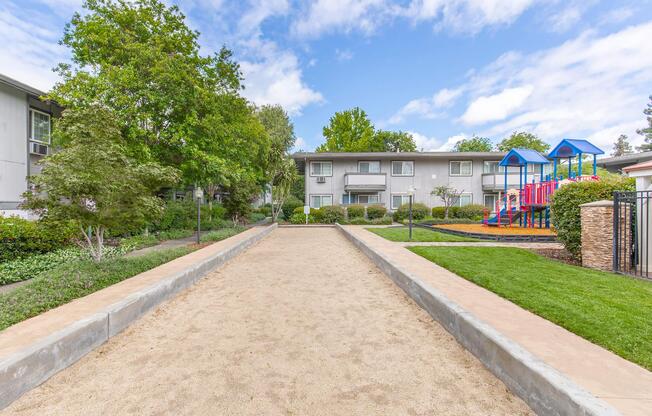 A pathway leading through a landscaped area, flanked by greenery and trees, with a playground featuring colorful equipment visible in the background. The sky is partly cloudy, and there are residential buildings on the left side of the image.