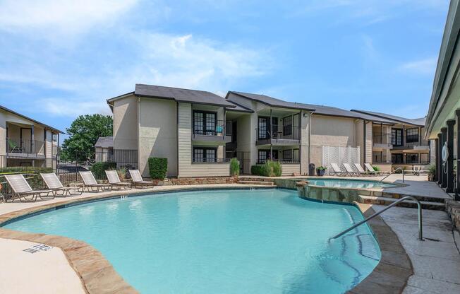 A tranquil swimming pool surrounded by lounge chairs and modern apartment buildings. The pool area features a stone edge and is flanked by greenery under a clear blue sky, creating a relaxing outdoor atmosphere for residents and guests.