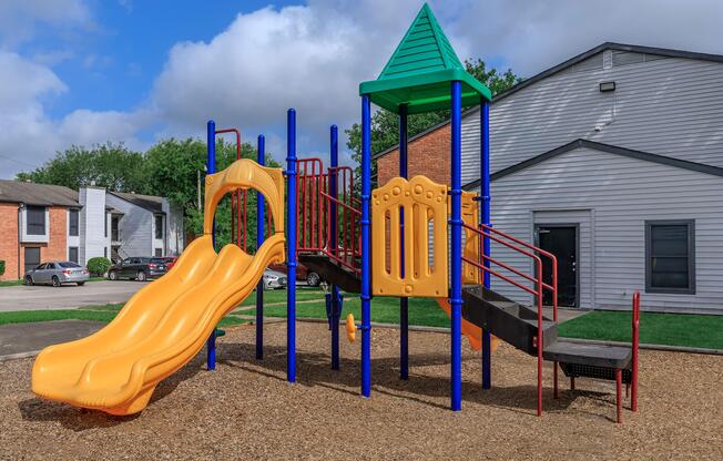 Colorful playground equipment featuring a yellow slide and a green triangular roof, surrounded by gravel and located near residential buildings.