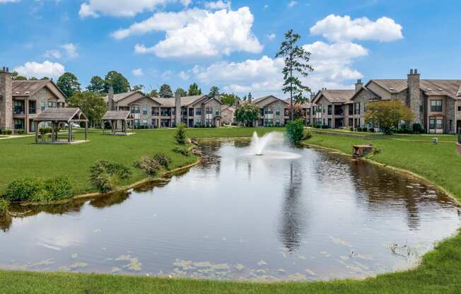 a pond with a fountain in the middle of a green field with houses