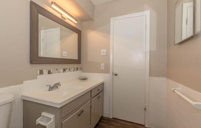 A modern bathroom featuring a double vanity with a sink, a large mirror above, and neutral-colored walls. There is a white toilet and a door leading to another room. The floor has a wood-like appearance, and a small decorative dish is placed on the counter. A light fixture above the mirror provides illumination.