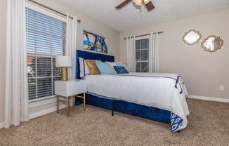 A beautifully decorated bedroom featuring a plush blue bed with white bedding, complemented by decorative pillows. Natural light streams through large windows, enhancing the cozy atmosphere. A stylish nightstand with a lamp and decorative art on the wall add elegance, while two decorative mirrors reflect the room's charm.