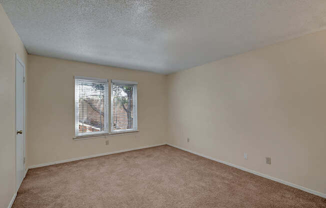 A bright neutral bedroom here at Summertree Place featuring soft carpet flooring, light beige walls, a textured ceiling, a single window with white blinds providing natural light, and a clean open layout that offers flexible space for bedroom furniture and personal touches.
