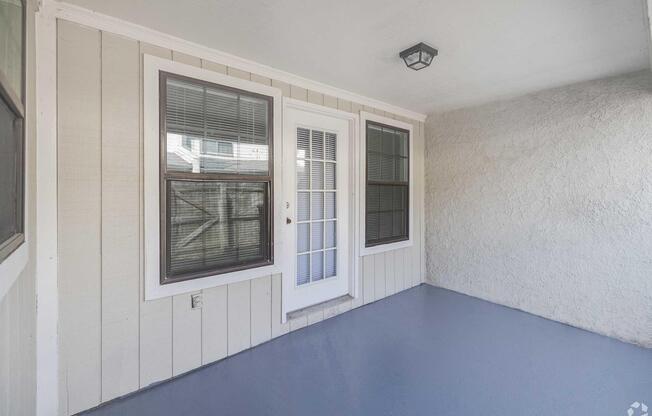 An enclosed porch featuring two large windows and a white door in the center. The floor is painted gray, and the walls are a light beige with textured finish. A ceiling light fixture is visible. The space is well-lit and offers a cozy outdoor view.
