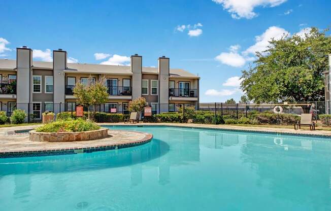 Here at High Plains, a bright outdoor swimming pool with clear blue water sits in a landscaped courtyard, framed by mature trees, fencing, and surrounding apartment buildings with balconies under a sunny blue sky.