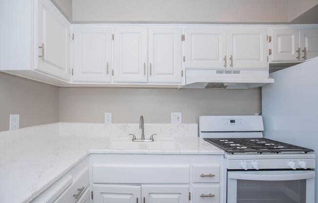 A modern kitchen featuring white cabinetry, a marble countertop, a stainless steel sink, and a gas stove. The kitchen also includes a fridge and a beige wall background, creating a clean and bright aesthetic.