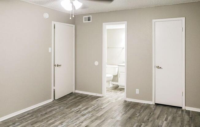 A minimalist room featuring light gray walls and wood-like flooring. Two closed doors are visible on the left and right, leading to separate spaces. The door on the left possibly leads to a bathroom, which has a visible light reflecting off a white wall. A ceiling fan is mounted above.