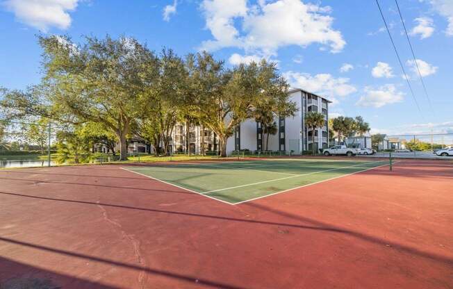 A tennis court with a red surface and white lines, surrounded by trees and a building in the background.