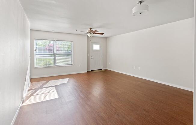 A spacious living room with wood-style flooring, large windows with natural light, and a ceiling fan at Muskego School Apartments