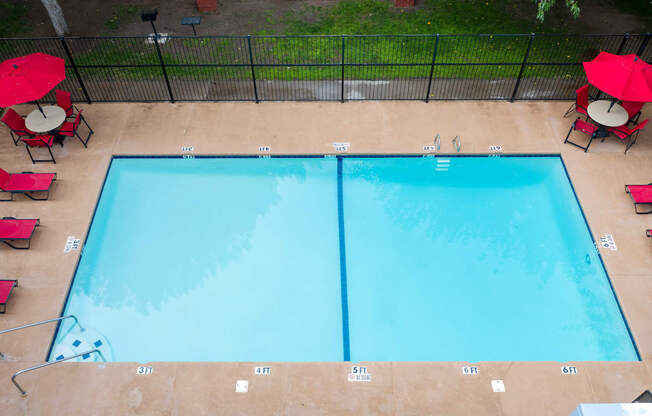 A rectangular pool with a blue tint is surrounded by a black fence and has red umbrellas on the side.