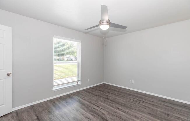 Empty room with light gray walls, a ceiling fan, and a window with blinds. The flooring is a light wood finish. Natural light is coming through the window, and the space appears clean and well-maintained, with no furniture present.