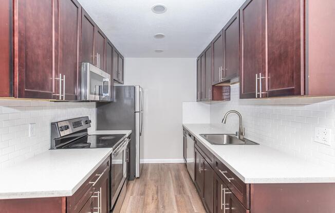 Modern kitchen featuring dark wooden cabinets, stainless steel appliances including a microwave, oven, and refrigerator. The countertops are smooth and light-colored, with a sleek sink and faucet. The flooring is wood-like, creating a warm ambiance in the space.