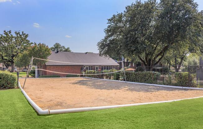 A sandy volleyball court surrounded by greenery, with a house visible in the background. The court features a net and is enclosed by a fence, under a clear blue sky. The area is well-maintained with grass in the foreground and trees providing shade.