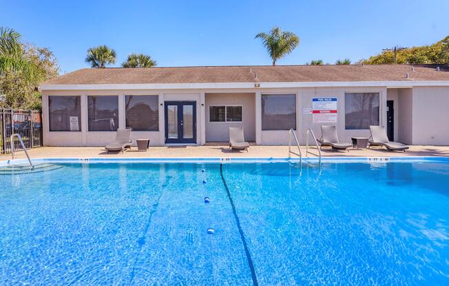 A clear blue swimming pool in front of a building with large windows. Several lounge chairs are placed around the pool area. Palm trees are visible in the background under a bright blue sky. The building features a door leading to an indoor space, and there is signage displayed nearby.