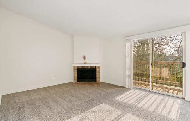 A large carpeted living room with white walls, a fireplace, and sliding glass doors to a private balcony with nature views.