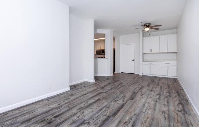 Spacious, modern open-concept living area featuring light-colored walls, laminate flooring, and a ceiling fan. The kitchen is partially visible with white cabinetry and stainless steel appliances. Natural light fills the room, creating a welcoming atmosphere.