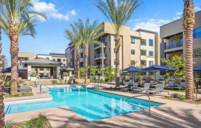A sunlit resort-style pool here at Reflect at Dobson Ranch showcasing a clear rectangular swimming pool with built-in steps and handrails, surrounded by tall palm trees, modern apartment buildings, landscaped greenery, and rows of lounge chairs with navy umbrellas on a spacious concrete deck, creating an inviting and relaxed community setting.