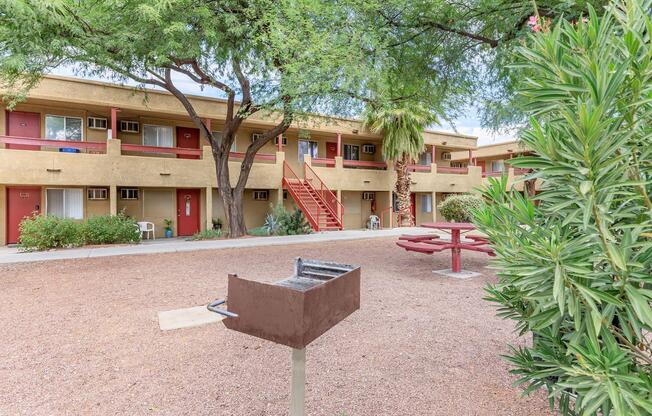 A courtyard view of a motel with two-story beige buildings, red doors, and balconies. There is a picnic area with a red table and a barbecue grill. Surrounding greenery includes shrubs and trees, providing a shaded and inviting atmosphere. The ground is covered in gravel.