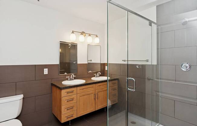 A bathroom with a white toilet, a wooden vanity with two sinks, and a glass shower stall.