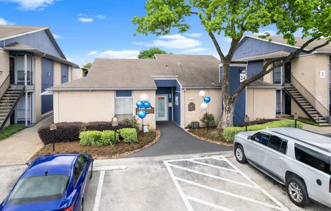A blue car is parked in a parking lot in front of a building with a tree and a white van.