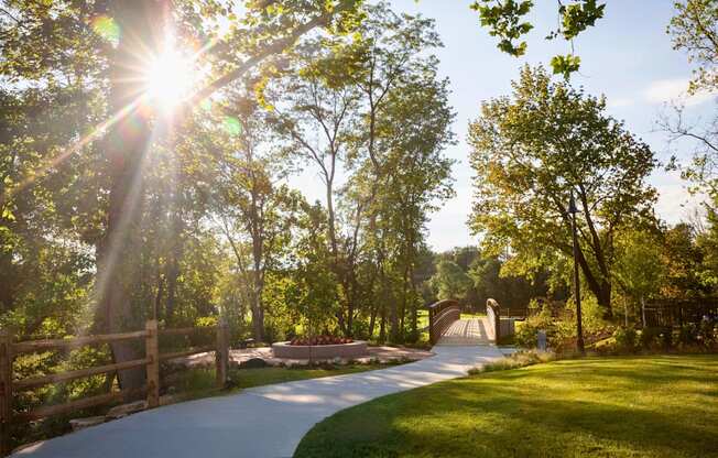 A beautiful trail with the sun shining through large trees and well-maintained grass leading to a bridge.