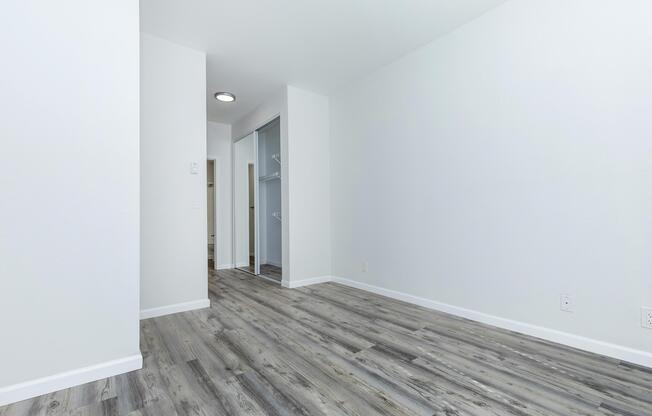 A spacious, empty room with light-colored walls and wooden flooring. The room features a closet area and an adjacent doorway leading to another room. Natural light illuminates the space, creating a bright and airy atmosphere.