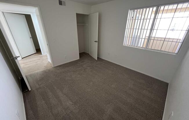 A vacant room with beige carpet, featuring a window with blinds and a closet with an open door. Another door leads to an adjoining room. The walls are painted white, and the space is well-lit from natural light.
