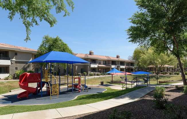 A playground with a red slide and blue umbrellas is surrounded by apartment buildings.